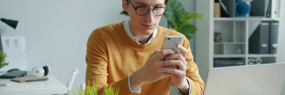 mężczyzna w okularach patrzący w telefon za biurkiem
